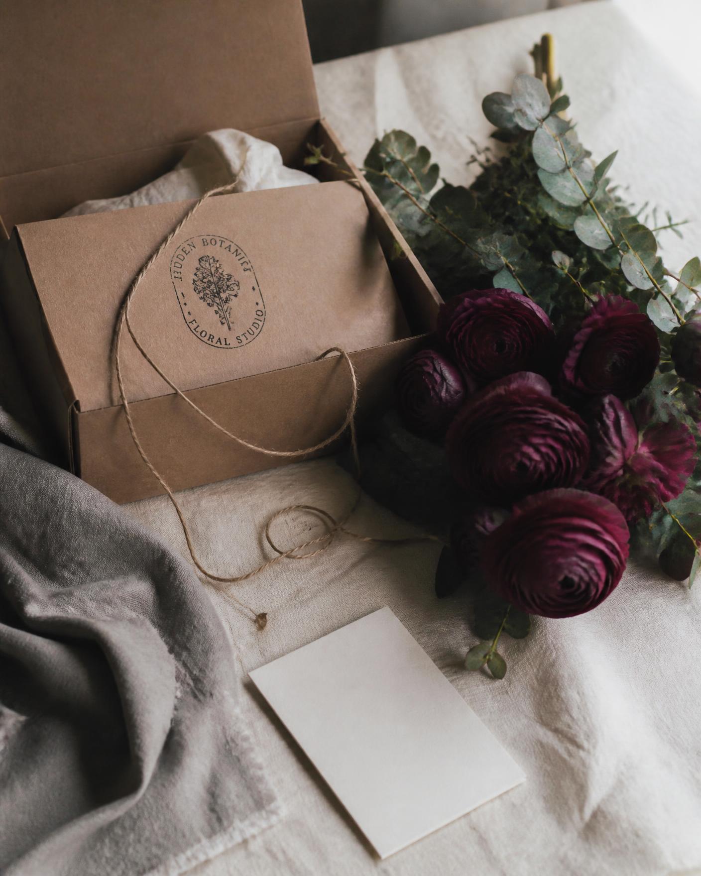 An open craft mailer with an ink-stamped inner box, deep berry ranunculus and eucalyptus on warm linen